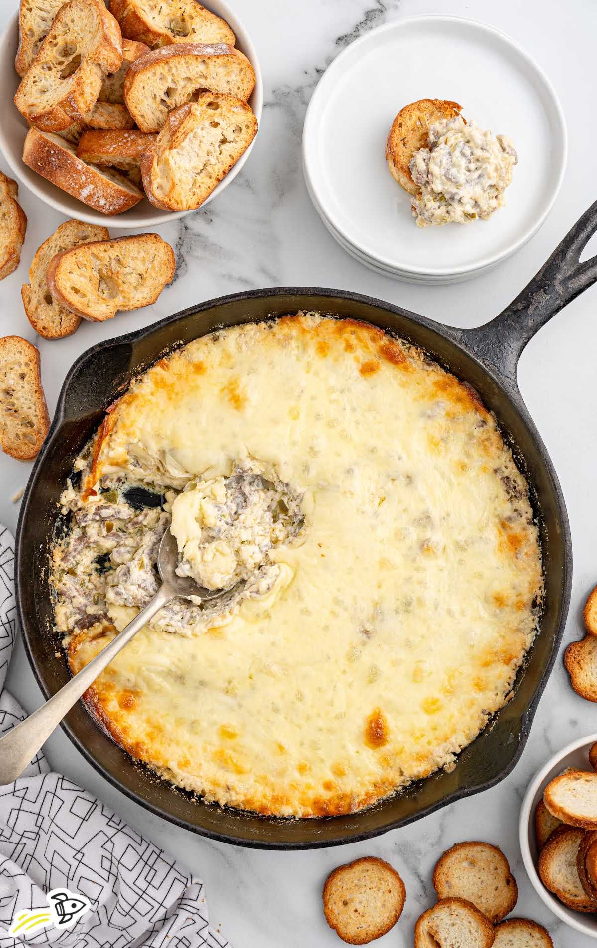 A top-down view of a skillet filled with Philly cheesesteak dip, with a serving spoon revealing the creamy filling, surrounded by slices of toasted bread.