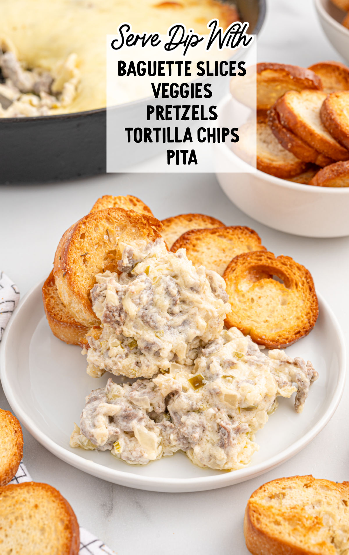 A plate of Philly Cheesesteak Dip with toasted baguette slices, featuring creamy melted cheese, seasoned steak, diced onions, and peppers. A cast iron skillet with the remaining dip is in the background.
