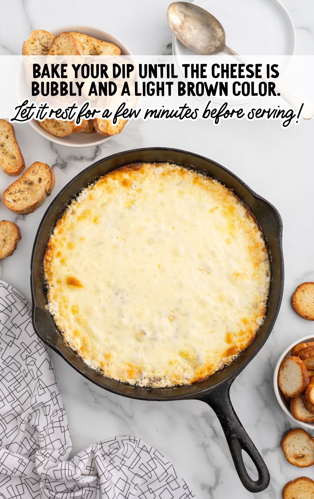 A cast iron skillet filled with baked Philly Cheesesteak Dip, topped with melted cheese that is bubbly and lightly browned. Toasted baguette slices are placed around the skillet, ready for dipping.