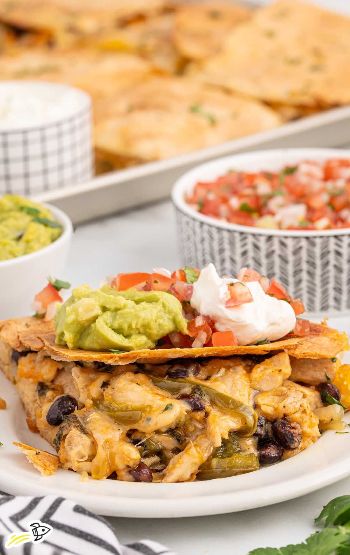 A serving of chicken sheet pan quesadilla topped with guacamole, sour cream, and pico de gallo on a white plate, with more quesadillas and dips in the background.