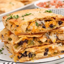 A plate stacked with cheesy chicken sheet pan quesadilla slices garnished with fresh cilantro, with a tray of quesadillas and pico de gallo in the background.