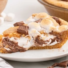 A whole s'mores pie topped with toasted marshmallows and Hershey's chocolate pieces, with a slice removed to reveal the gooey layers.