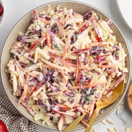 Overhead image of prepared apple slaw in a bowl with gold utensils and colorful shredded ingredients visible.