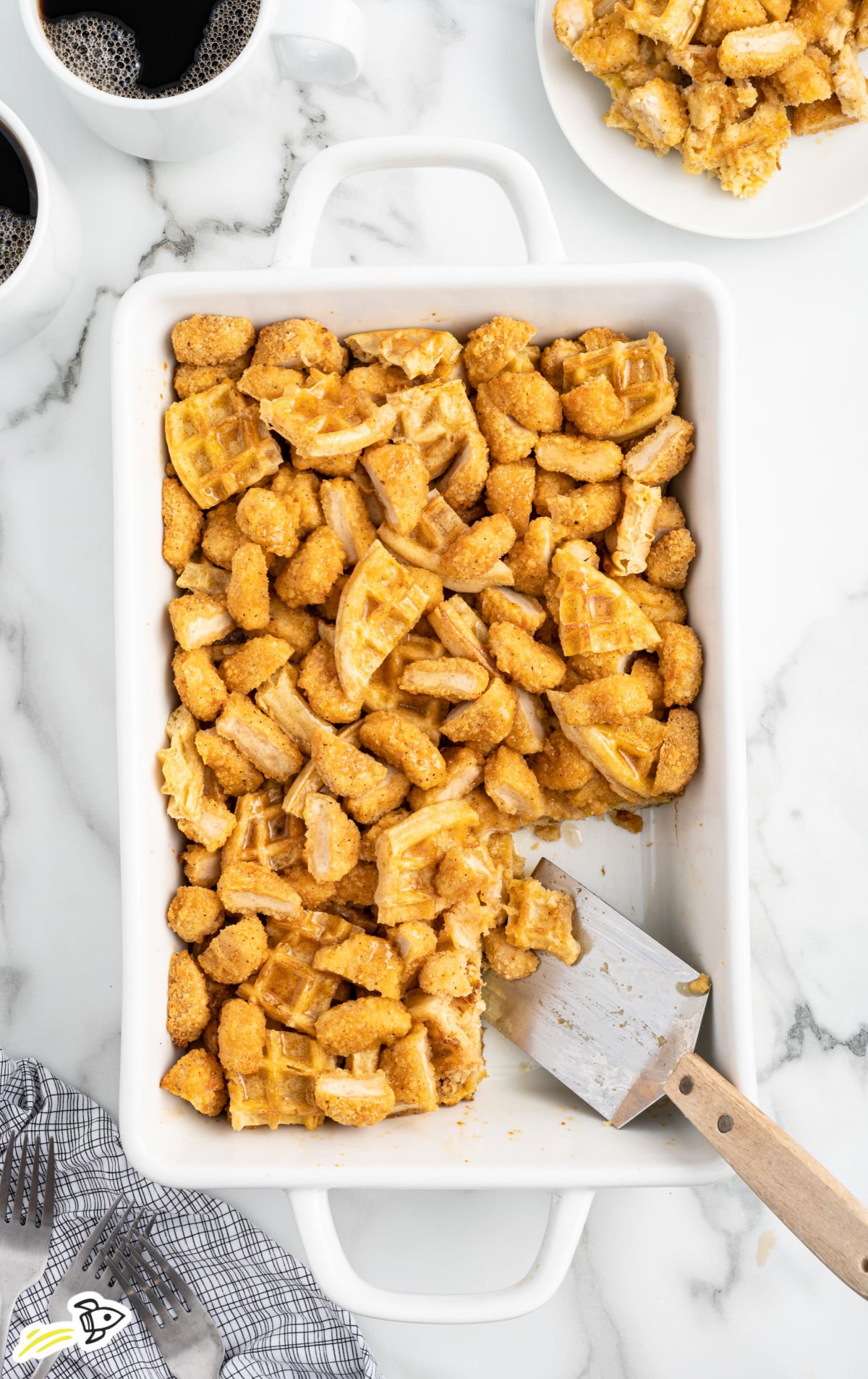 Overhead view of a white baking dish filled with golden brown chicken and waffle casserole, with coffee mugs and a plated serving nearby.