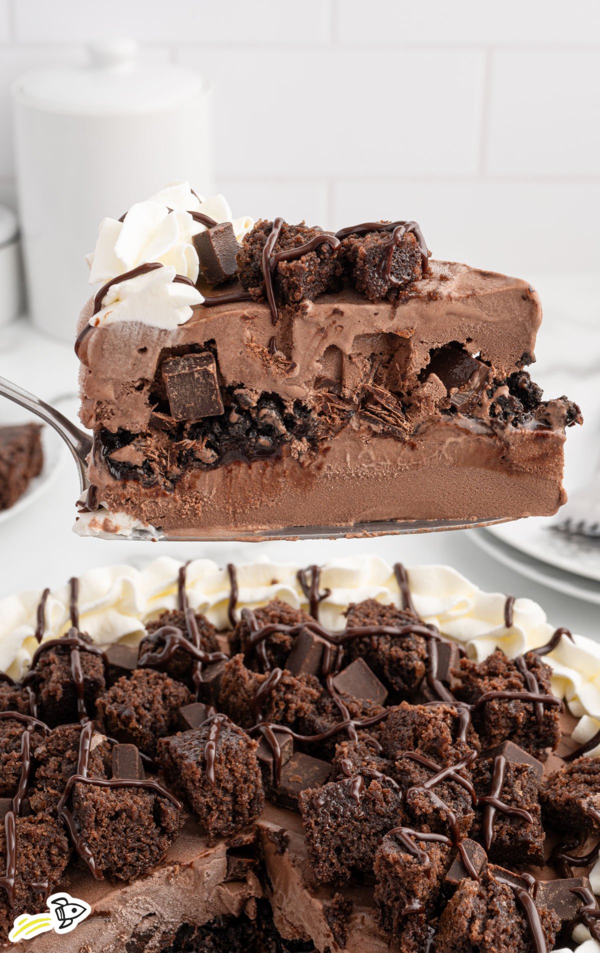 Close-up of a single slice being lifted from the whole cake, showing detailed layers and textures including brownie pieces, whipped cream, and cookie crunch.