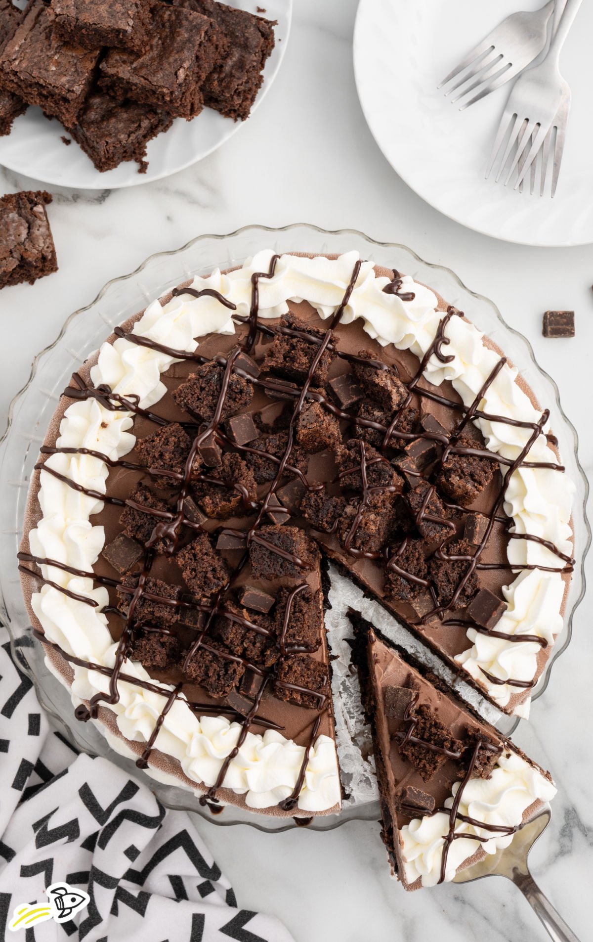 Overhead view of a sliced Dairy Queen copycat chocolate ice cream cake topped with whipped cream, brownie chunks, chocolate pieces, and chocolate drizzle, with one slice being lifted from the cake.