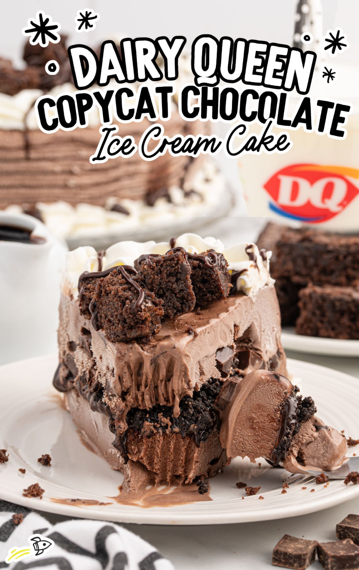 Close-up of a slice of Dairy Queen copycat chocolate ice cream cake on a white plate, showing distinct layers of chocolate ice cream, fudge, crushed cookie crunch, and topped with whipped cream, brownie chunks, and chocolate drizzle. A glass of milk with the DQ logo is in the background.