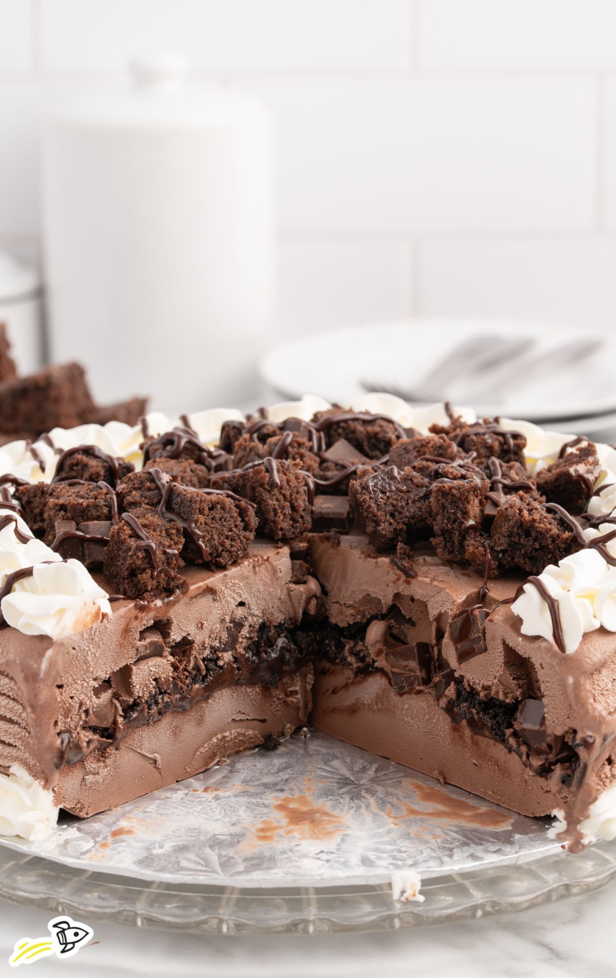 Close-up of a partially sliced Dairy Queen copycat chocolate ice cream cake on a glass platter, showing distinct layers of chocolate ice cream, fudge, crushed cookies, and chocolate chunks, topped with whipped cream and brownie pieces drizzled with chocolate syrup.