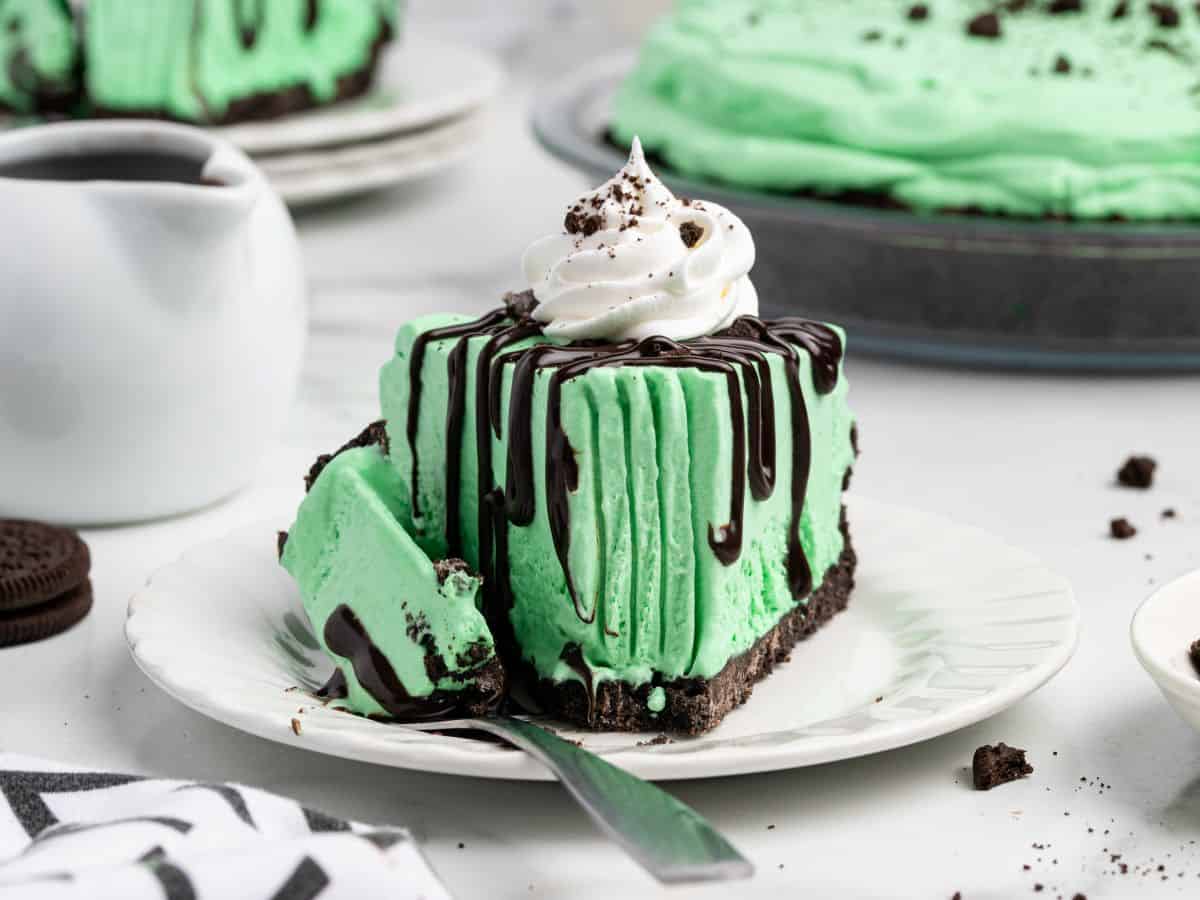 A slice of Grasshopper Pie—a mint chocolate ice cream pie with whipped cream and chocolate drizzle—sits on a plate with a fork. In the background, a whole Grasshopper Pie, a cup, and cookie crumbs complete the tempting scene.
