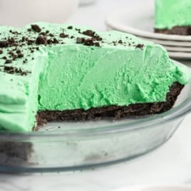 A mint green Grasshopper Pie with a chocolate cookie crust sits in a glass pie dish. One slice is missing, revealing the creamy texture inside. Crumbled chocolate cookies are sprinkled on top.