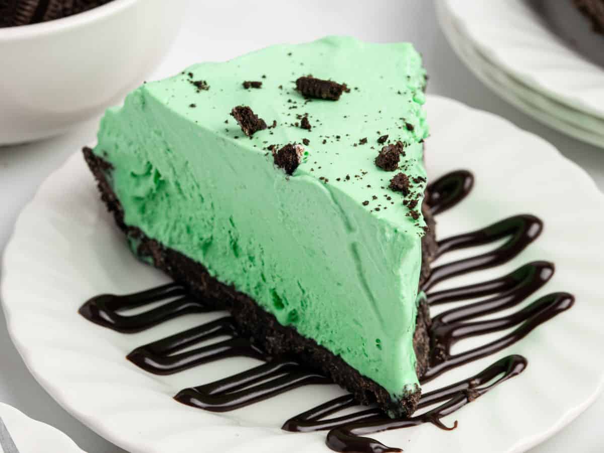 A slice of Grasshopper Pie with a chocolate cookie crust, topped with cookie crumbs, sits on a white plate drizzled with chocolate syrup.