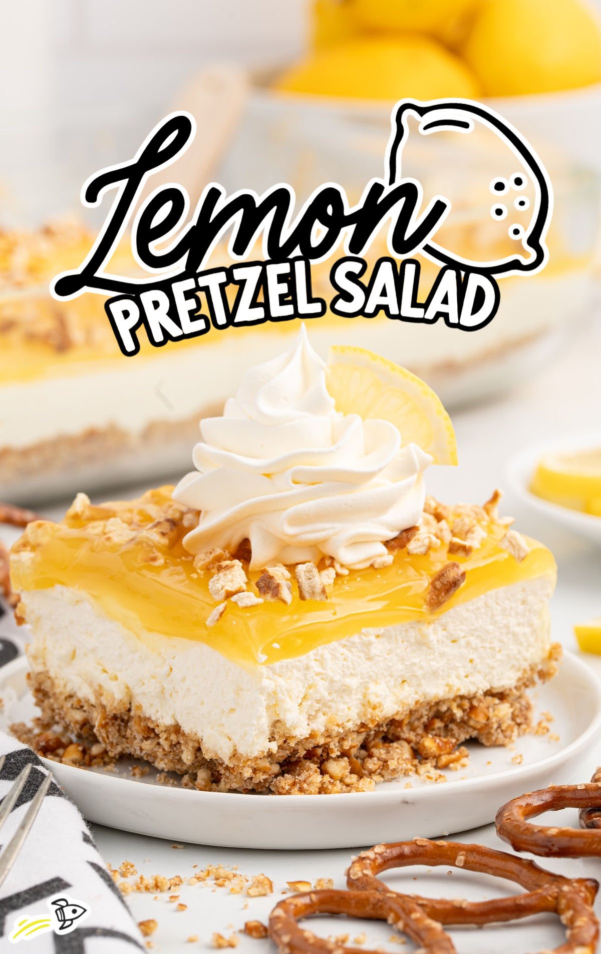 A slice of lemon pretzel salad on a white plate, featuring a crunchy pretzel crust, creamy cheesecake-style filling, and a glossy lemon topping. Garnished with whipped cream, crushed pretzels, and a lemon wedge. Whole pretzels and lemon slices are scattered nearby, with the full dessert in a glass dish in the background.