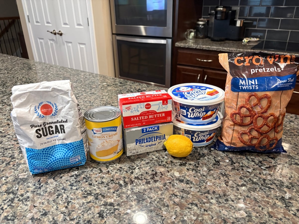 Various ingredients for Lemon Pretzel Salad are lined up on a kitchen counter, including granulated sugar, a can of lemon creme, cream cheese, butter, lemon, Cool Whip containers, and a bag of mini pretzel twists.