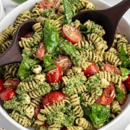 Overhead view of a large white bowl filled with pesto pasta salad, garnished with fresh basil and served with wooden utensils.