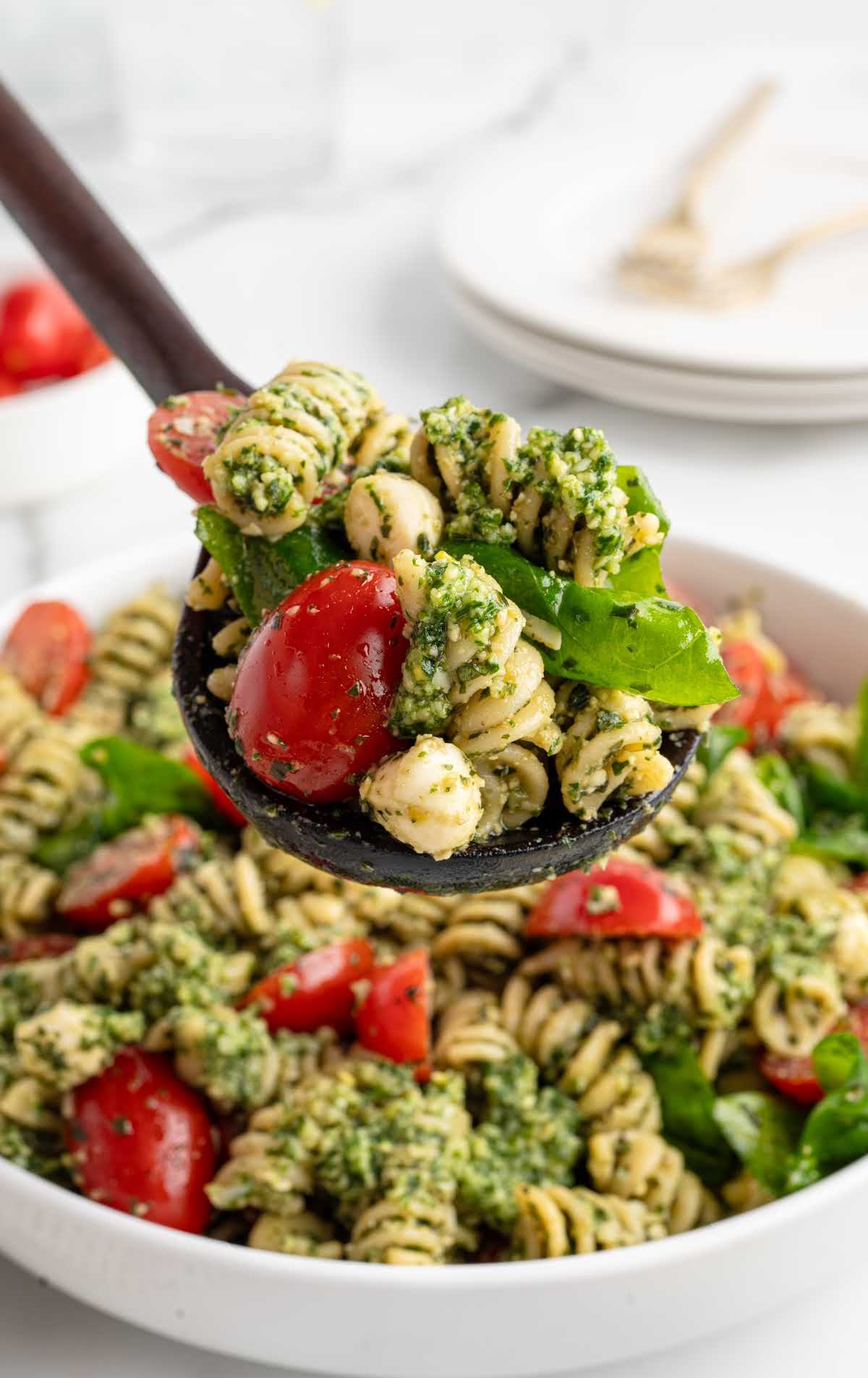 Wooden spoon lifting a serving of pesto pasta salad with cherry tomatoes, mozzarella balls, and spinach.