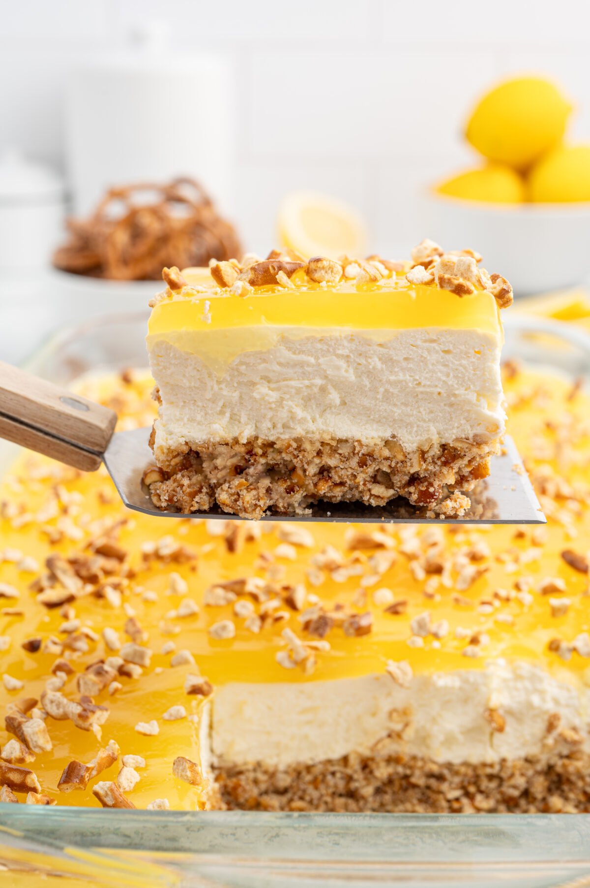 A close-up of a spatula lifting a slice of lemon pretzel salad from a glass baking dish. The dessert has a crunchy pretzel crust, creamy filling, and a glossy lemon topping, garnished with chopped pretzels. The remaining dessert and whole lemons are visible in the background.