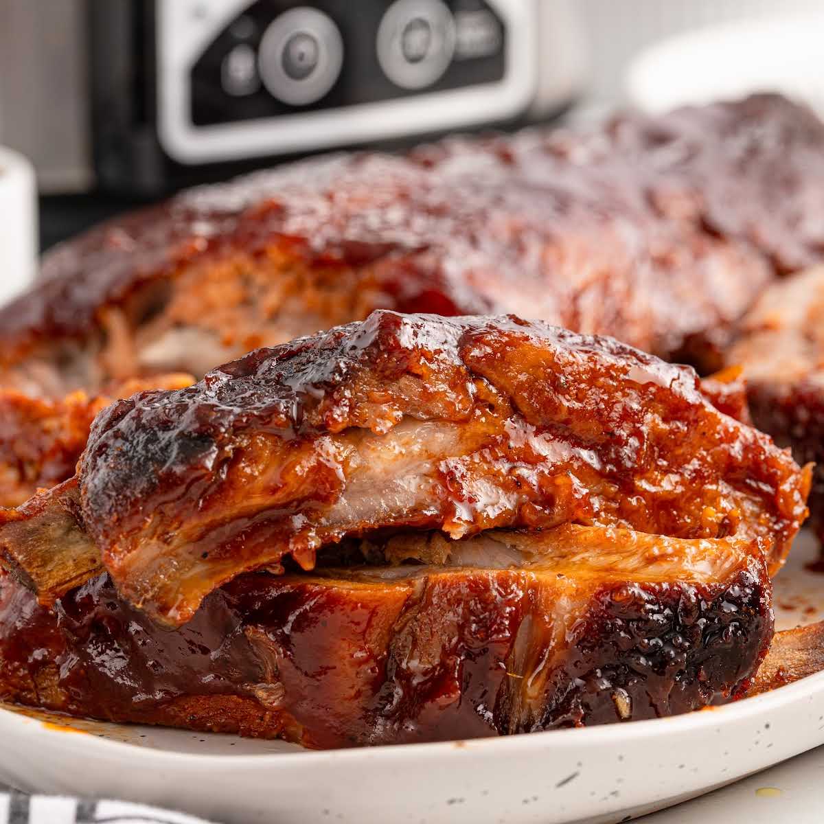 Close-up of sliced slow cooker baby back ribs on a white plate, glistening with BBQ sauce, with a slow cooker blurred in the background.