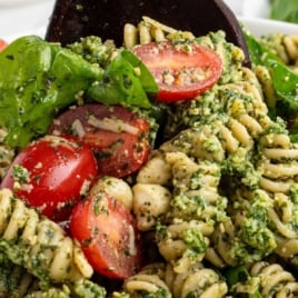 Overhead shot of a vibrant pesto pasta salad with rotini, cherry tomatoes, spinach, and mozzarella in a white bowl with bold text overlay.