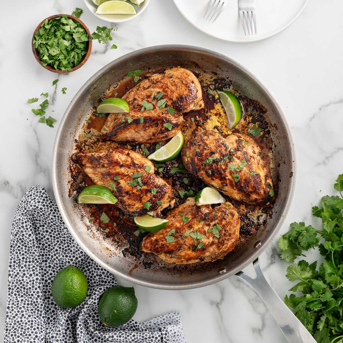 Square image of grilled cilantro lime chicken breasts in a skillet, surrounded by lime wedges and fresh cilantro.