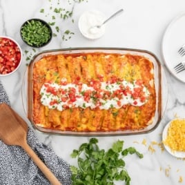 Overhead shot of cream cheese chicken enchiladas surrounded by ingredients like shredded cheese, chopped tomatoes, and fresh cilantro, styled for a recipe card.