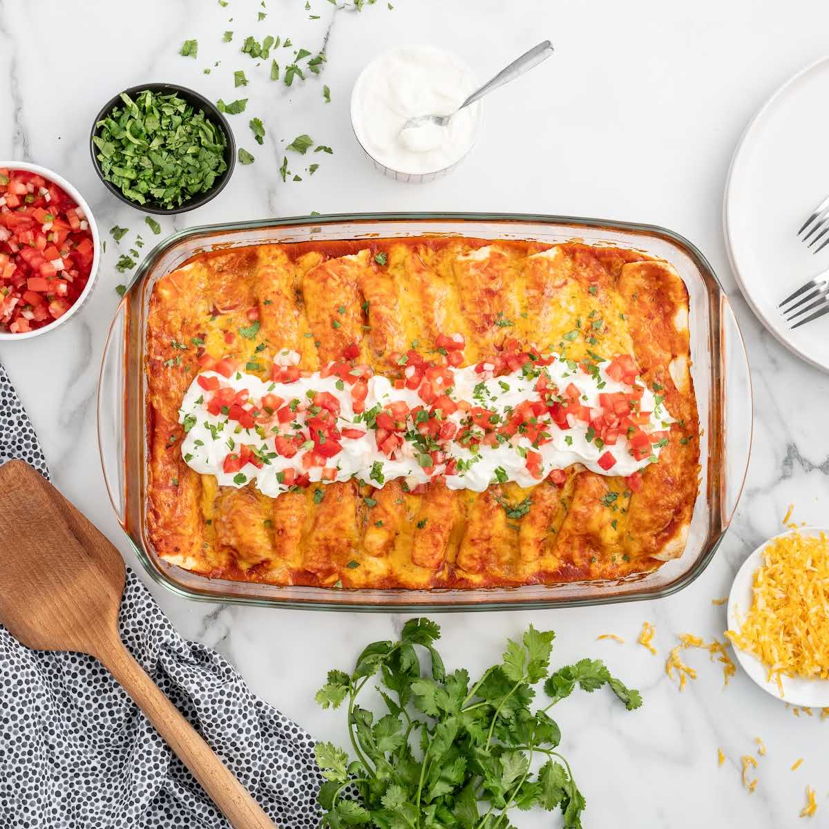 Overhead shot of cream cheese chicken enchiladas surrounded by ingredients like shredded cheese, chopped tomatoes, and fresh cilantro, styled for a recipe card.