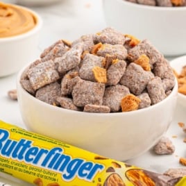 Bowl of Butterfinger muddy buddies next to a whole Butterfinger bar, with peanut butter and candy bowls in the background.