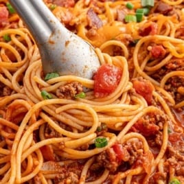 Large skillet of cowboy spaghetti with rich tomato sauce, ground beef, and noodles, being tossed with metal tongs.