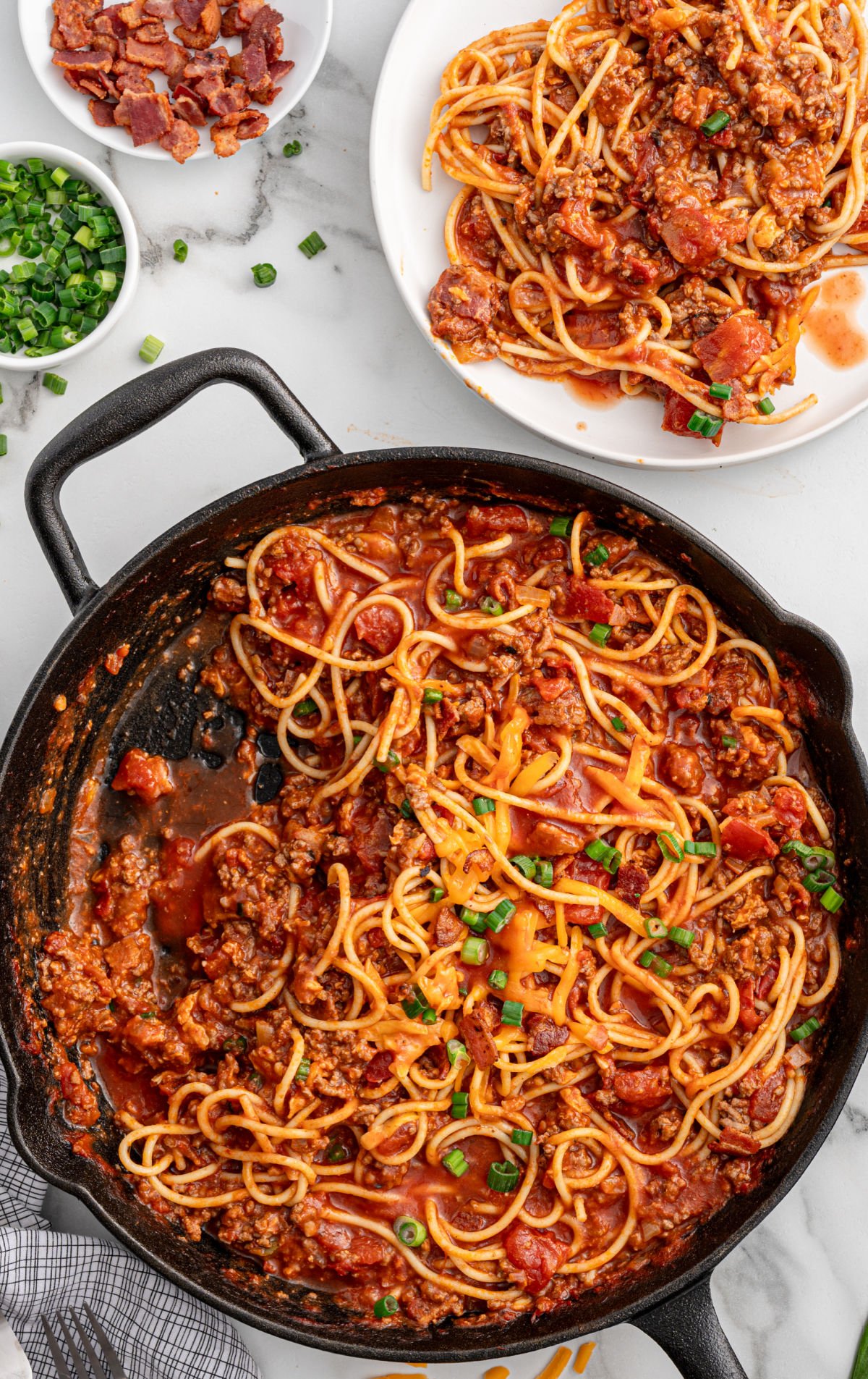 Overhead view of a skillet filled with cowboy spaghetti, melted cheese in the center, surrounded by saucy spaghetti noodles, ground beef, and diced tomatoes, garnished with green onions.