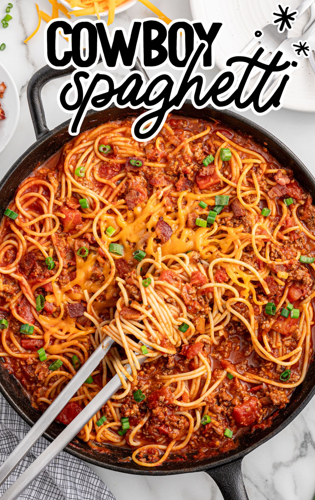 Overhead view of a skillet filled with cowboy spaghetti, melted cheese in the center, surrounded by saucy spaghetti noodles, ground beef, and diced tomatoes, garnished with green onions.