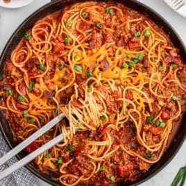 Overhead view of a skillet filled with cowboy spaghetti, melted cheese in the center, surrounded by saucy spaghetti noodles, ground beef, and diced tomatoes, garnished with green onions.