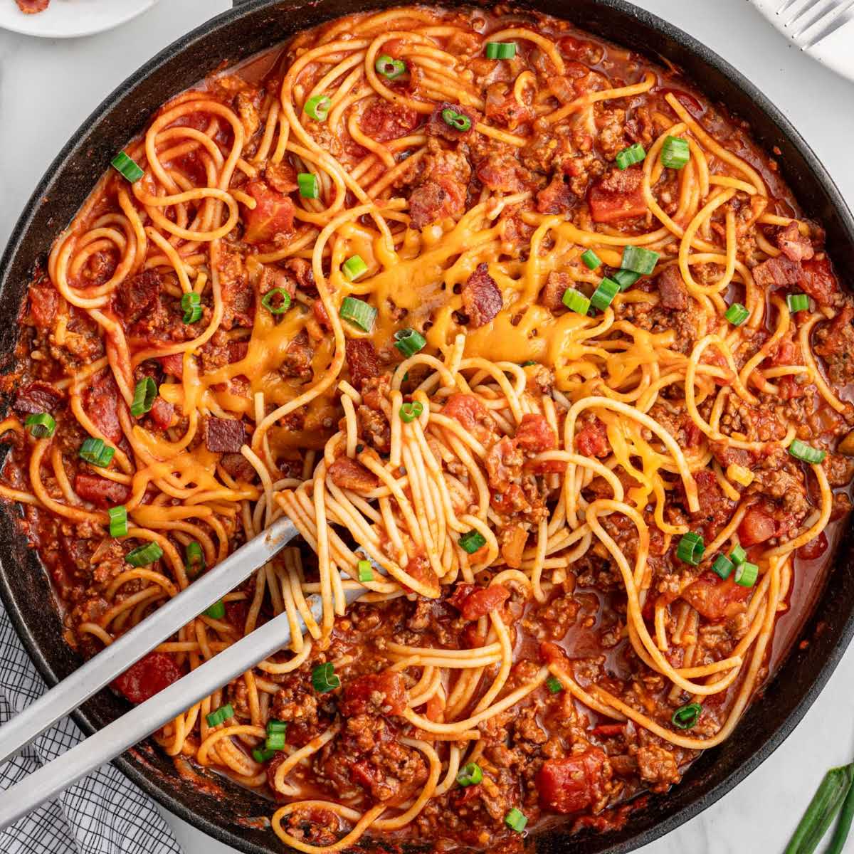 Overhead view of a skillet filled with cowboy spaghetti, melted cheese in the center, surrounded by saucy spaghetti noodles, ground beef, and diced tomatoes, garnished with green onions.