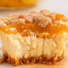 Mini peach cobbler cheesecake on a white plate with a crumbly brown sugar topping, juicy peach slices, and creamy cheesecake center, with syrup pooling on the plate.