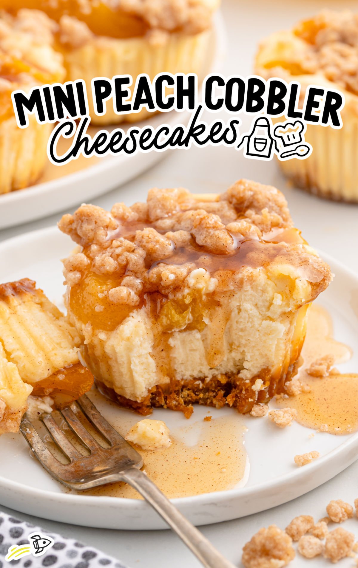 Mini peach cobbler cheesecake with a bite taken out, showing creamy filling and crumbly topping, with syrup pooling on the plate. Text overlay reads "Mini Peach Cobbler Cheesecakes".