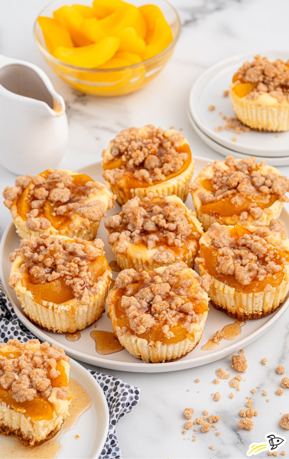 A serving plate piled with mini peach cobbler cheesecakes next to a bowl of canned peach slices and a small white syrup pitcher.