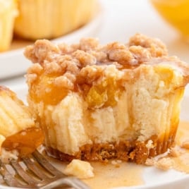 Single mini peach cobbler cheesecake on a fork, partially eaten to show creamy interior and peach filling, resting in a pool of sweet syrup.