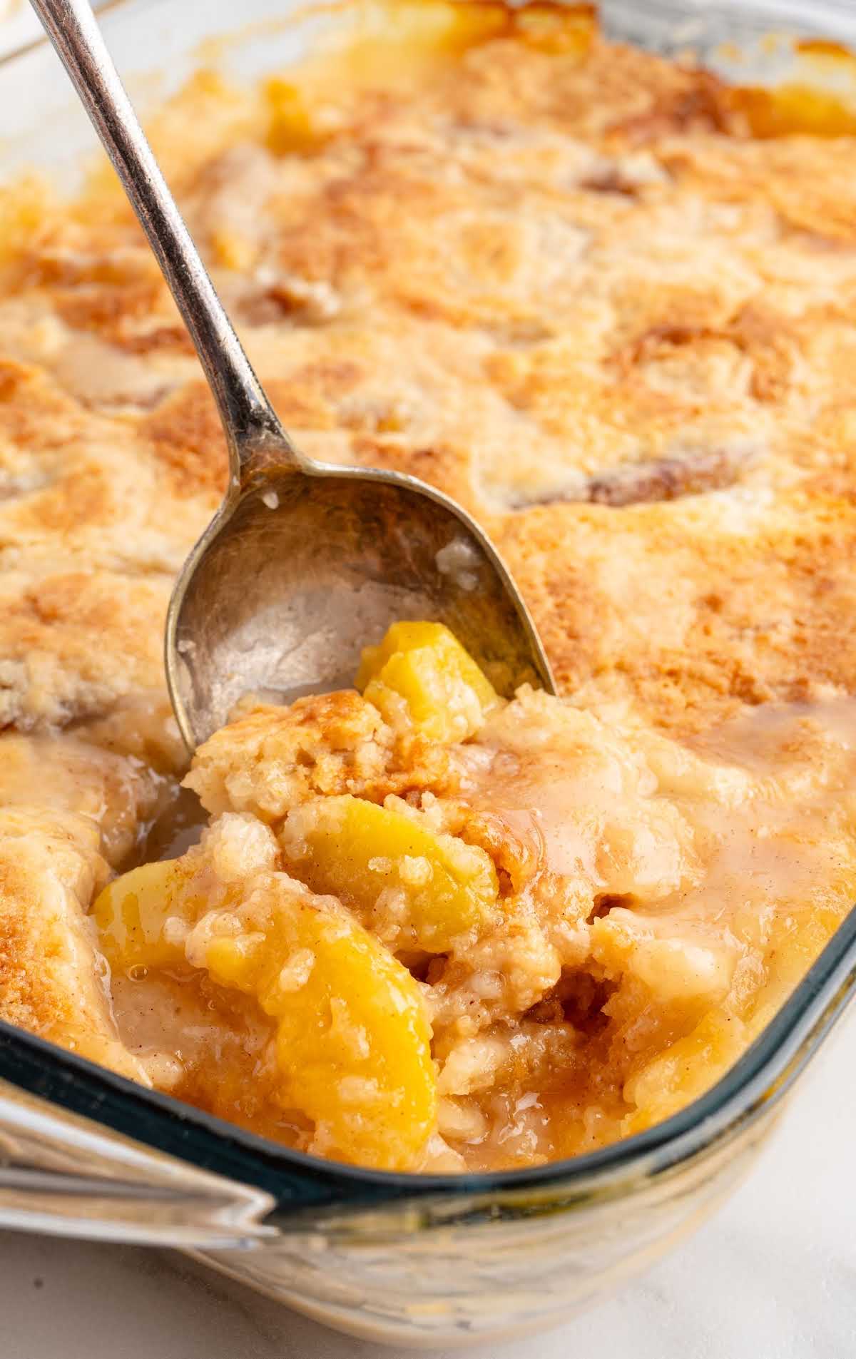 Glass baking dish filled with freshly baked peach dump cake, with a serving spoon scooping out a portion.