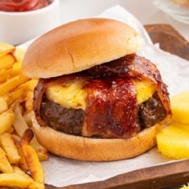 pineapple bacon burger with fries and pineapple slices, styled on a wooden tray with a white parchment paper.