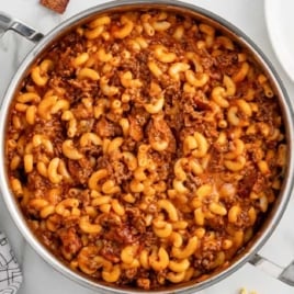 Overhead shot of cheesy bacon cheeseburger pasta in a stainless steel skillet with scattered cheese and bacon on a marble countertop.