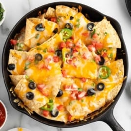 Skillet of cheesy nachos topped with black olives, jalapeños, tomatoes, and melted cheese, surrounded by fresh toppings.