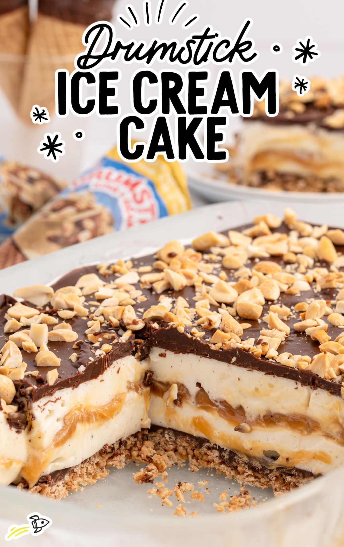 Drumstick ice cream cake with a crunchy sugar cone crust, creamy vanilla ice cream, caramel swirls, a thick layer of chocolate magic shell, and chopped peanuts on top. A slice is cut out to reveal the layered interior.