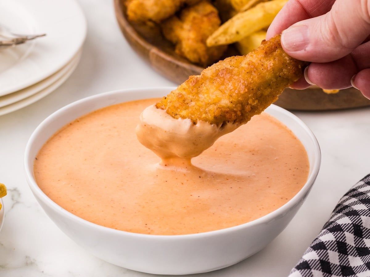 A hand dips a breaded chicken strip into a bowl of creamy, orange Red Robin Campfire Sauce, with more chicken strips and potato wedges on a plate in the background.