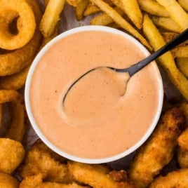 Bowl of creamy Red Robin campfire sauce surrounded by onion rings, fries, and chicken tenders on a tray.