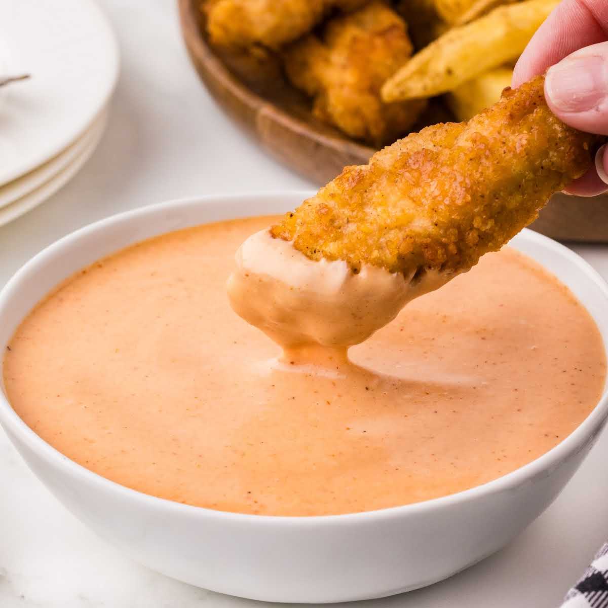 Crispy chicken strip being dipped into a bowl of creamy campfire sauce, with fries and tenders in the background.