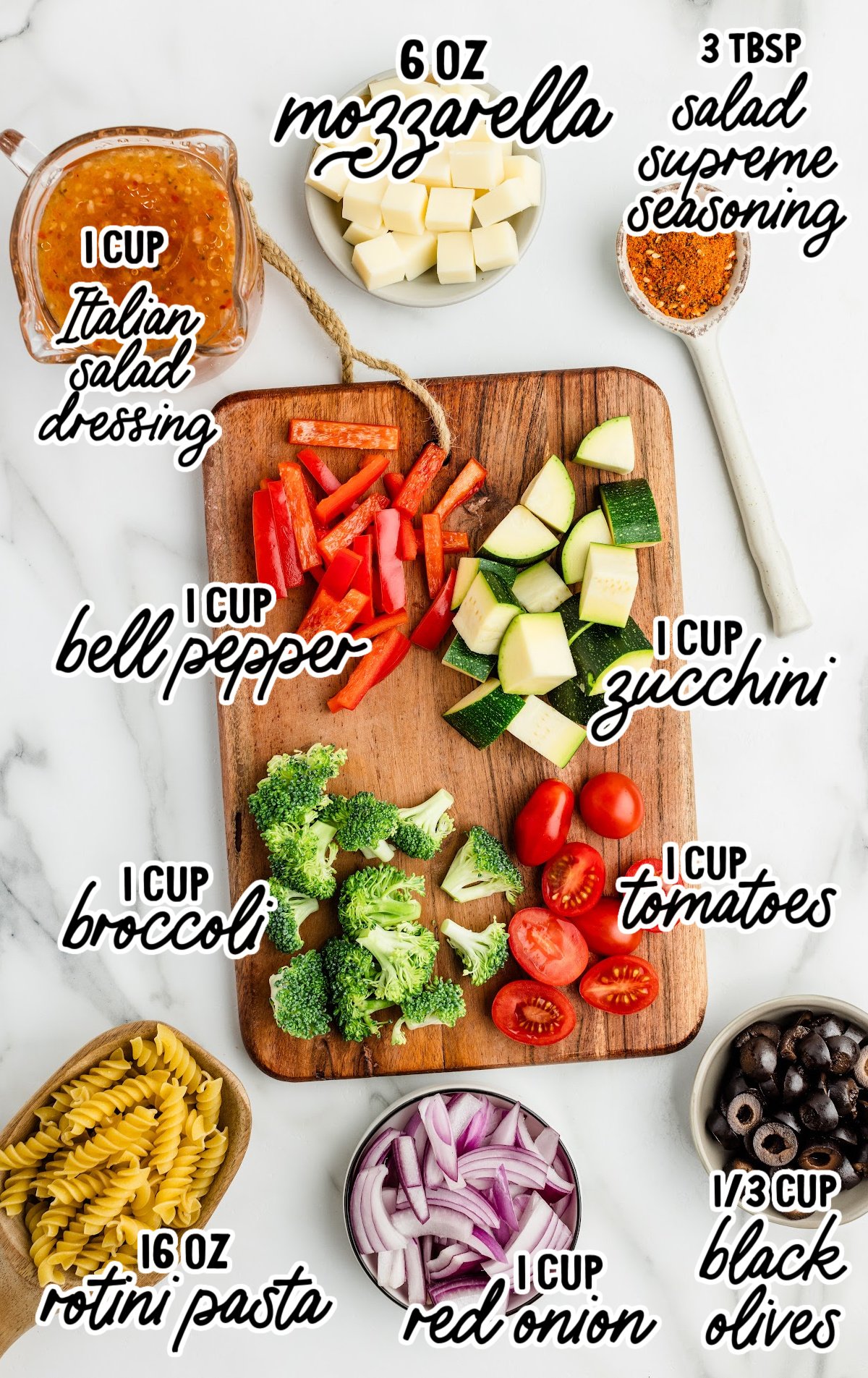 Flat lay of pasta salad ingredients arranged on a white marble surface. A wooden cutting board holds chopped red bell pepper, zucchini, broccoli, and grape tomatoes. Surrounding the board are labeled bowls and containers with 1 cup Italian salad dressing, 6 oz mozzarella cubes, 3 tablespoons Salad Supreme seasoning, 16 oz rotini pasta, 1 cup red onion slices, ⅓ cup sliced black olives, and additional bowls of the chopped vegetables. Each ingredient is clearly labeled with handwritten-style text.