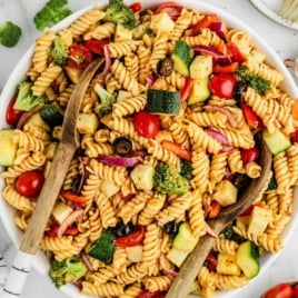 Top-down image of pasta salad with serving utensils, filled with fresh vegetables and tossed with dressing and seasoning