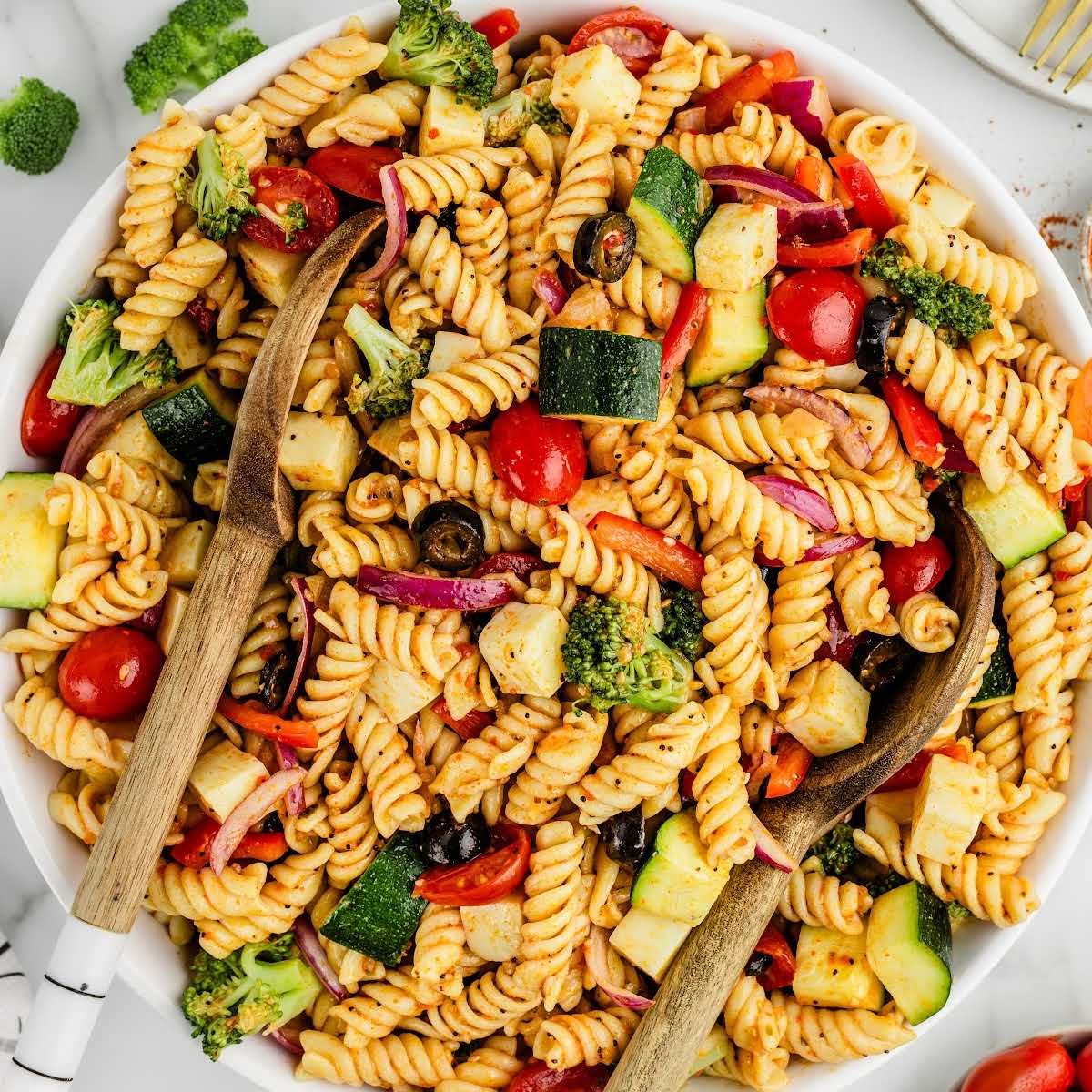 Top-down image of pasta salad with serving utensils, filled with fresh vegetables and tossed with dressing and seasoning
