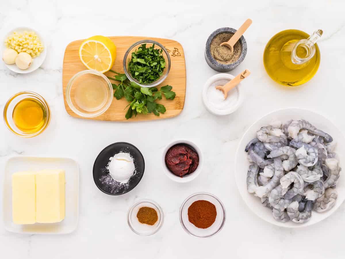 Assorted ingredients for Spicy Garlic Shrimp are displayed on a white surface, including raw shrimp, butter, olive oil, parsley, garlic, lemon, spices, tomato paste, salt, and pepper arranged in bowls and on a wooden board.