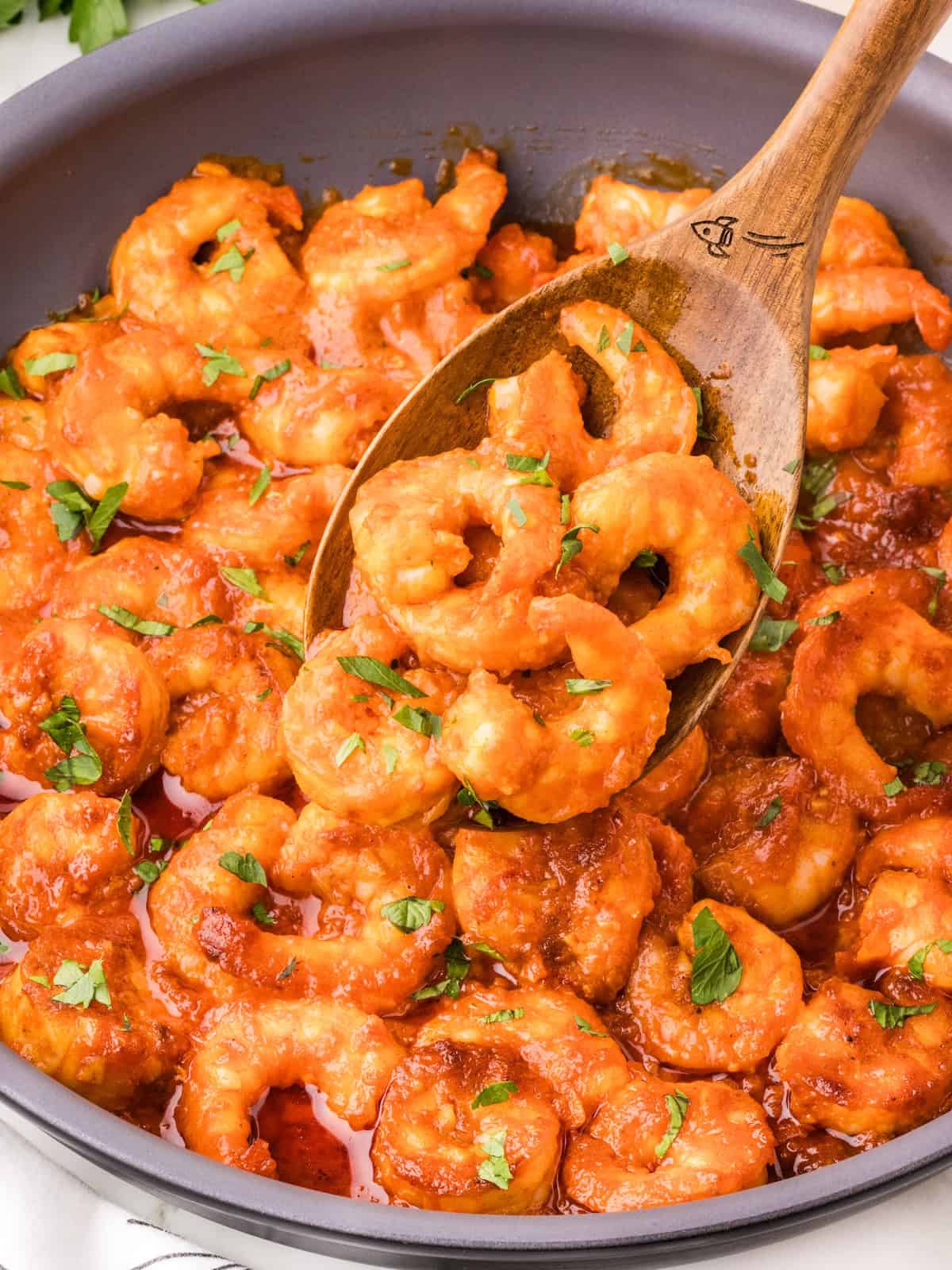 Spicy Garlic Shrimp cooked in a rich red tomato sauce, garnished with chopped parsley, being stirred with a wooden spoon in a pan.