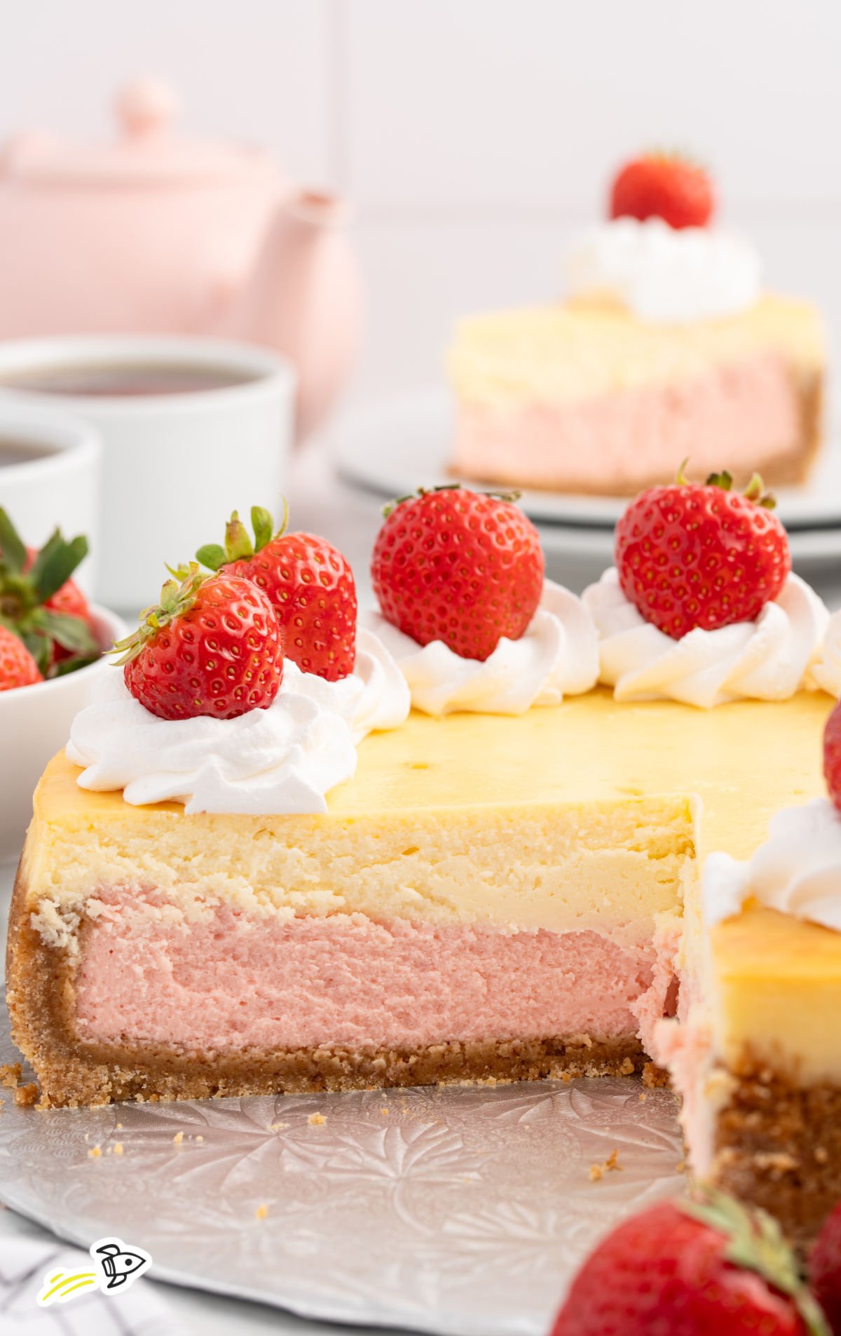 Strawberries and cream cheesecake on a cake stand with one slice removed, topped with whipped cream dollops and fresh strawberries, set against a tea-time backdrop with cups and a pink teapot.