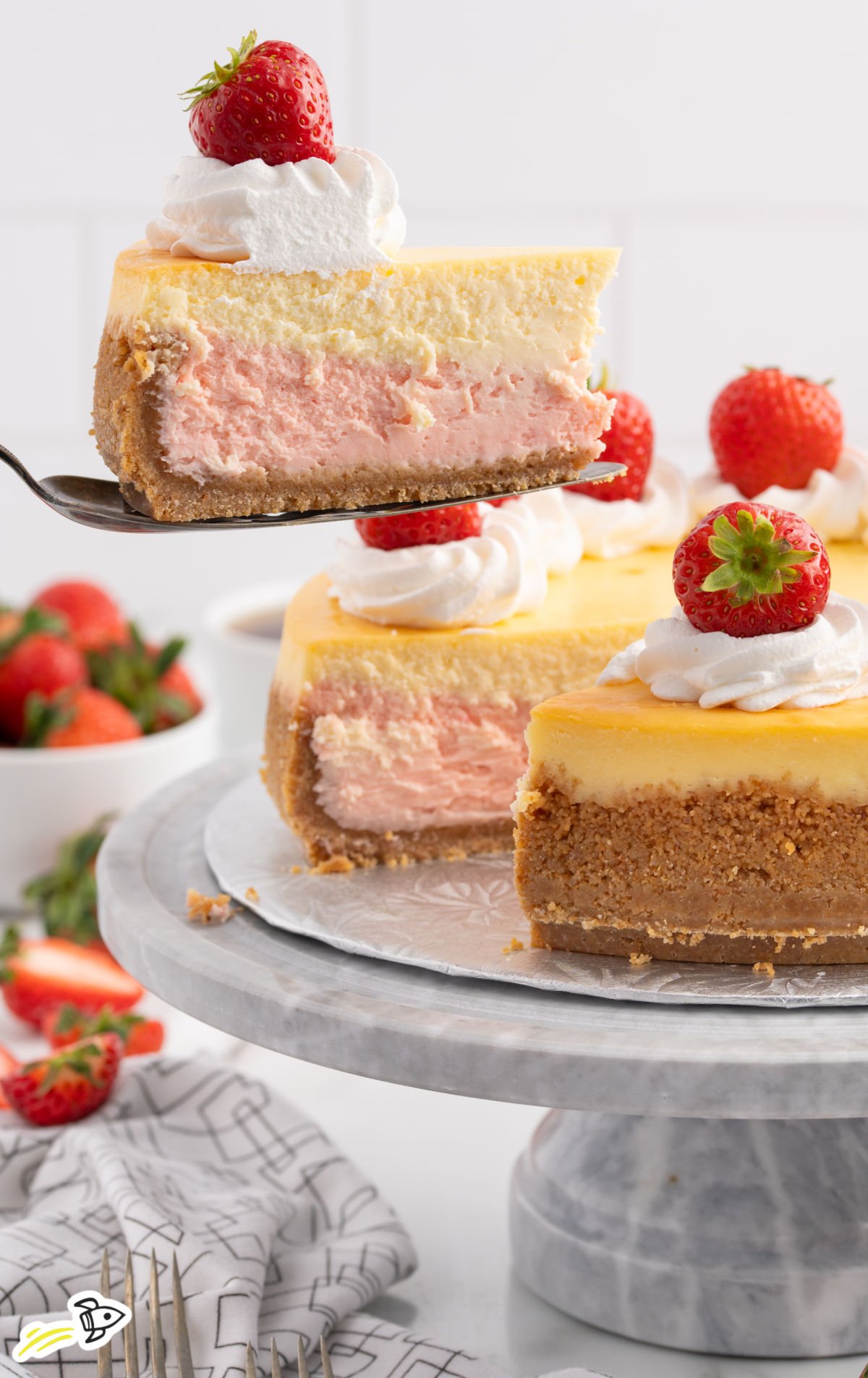 A slice of strawberries and cream cheesecake being lifted from a whole cheesecake on a marble cake stand. The slice shows distinct layers of pink strawberry and creamy vanilla cheesecake on a graham cracker crust, topped with whipped cream and a fresh strawberry.